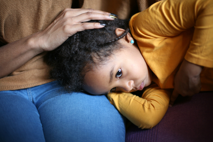 A young girl lays her head in her mother's lap. The mother rests her hand on the child's head.