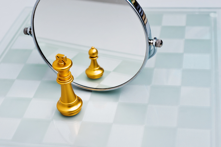 A golden king piece sits in front of a mirror. Its reflection shows a tiny pawn piece.
