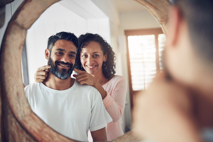 A wife encourages her husband to smile at himself in the mirror.