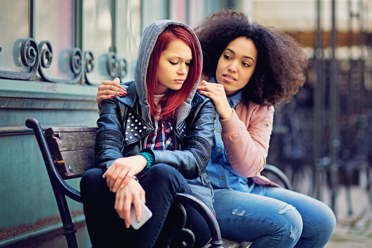 Two girls sit on a bench outside. One comforts the other after an upsetting phone call.