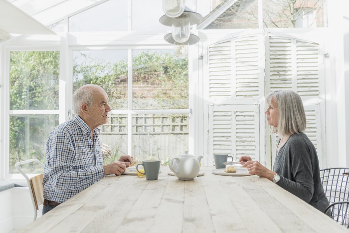 Older couple sits across from each other, eating in a conservatory.