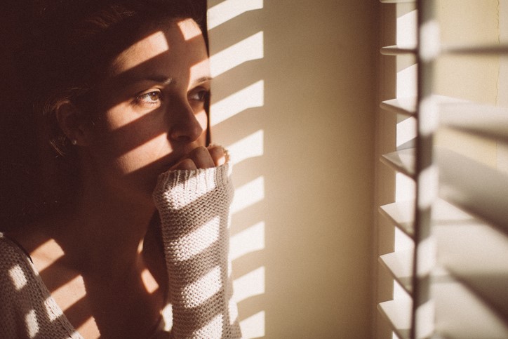 A young woman bites her nails as she stairs out her window. Sun shines in through half-open blinds.