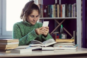 Woman sitting at desk, procrastinating with a smartphone next to open textbooks